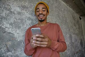Joyful beautiful brown eyed young dark skinned male with beard wearing pink sweater and mustard cap while posing over concrete background, keeping smartphone in hand and chatting with his friends photo