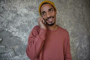 foto de alegre encantador oscuro desollado chico con barba vestido en casual ropa posando terminado hormigón pared mientras haciendo llamada con su móvil teléfono, mirando aparte con encantador sonrisa