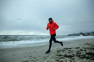 Full-length image of young active bearded male dressed in warm sporty clothes working out and running by seaside on cold gloomy weather. Sport and healthy lifestyle concept photo