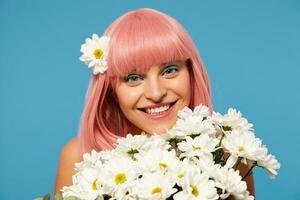 Studio photo of young pretty romantic pink haired woman with festive makeup posing in white flowers over blue background, looking positively at camera with charming smile