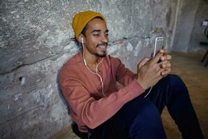 Horizontal shot of positive bearded dark skinned guy in pink sweater, blue pants, trousers and mustard cap sitting on wooden floor with smartphone in hands and listening to music with earphones photo