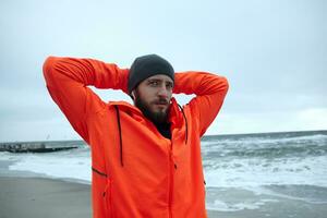 Close-up of young brunette bearded sportsman in black cap and warm orange coat standing over seaside on grey stormy weather and enjoying view after morning running session photo