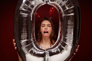 Close-up of young funny pretty female with wavy hairstyle fooling and making faces while posing over claret background with huge air number balloon. Holiday preparation and celebration concept photo