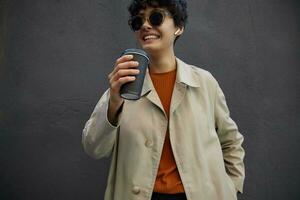 Happy lovely young dark haired woman with casual hairstyle wearing trendy outfit and stylish sunglasses while standing over black urban wall, keeping takeaway coffee in raised hand and smiling happily photo