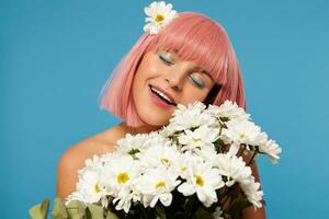 Romantic shot of young attractive pink haired female with colored makeup smiling pleasantly with closed eyes while posing in white camomiles over blue background photo