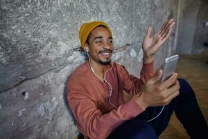 Pleased smiling young dark skinned male with beard raising palm and smiling happily while sitting over wooden floor in casual clothes, making video call with his smartphone photo