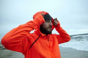 de cerca de joven hermoso barbado hombre participación elevado mano en capucha de su calentar atlético naranja Saco mientras respiración Fresco aire terminado playa, mirando adelante con calma cara foto