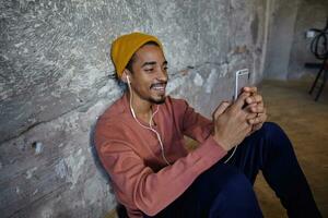 Positive pretty bearded dark skinned guy leaning on concrete wall while sitting on floor, wearing earphones and holding smartphone in raised hands, smiling while looking on screen photo