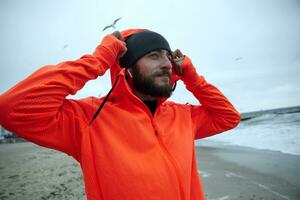 positivo joven morena barbado masculino en deportivo ropa escuchando a música en su auriculares y disfrutando Fresco clima terminado playa, siendo calma y sonriente ligeramente foto