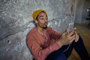 Indoor photo of pretty bearded dark skinned guy in pink sweater, blue pants, trousers and mustard cap leaning on concrete wall, making photo of himself with mobile phone and folding lips in air kiss