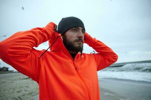 Outdoor shot of young pretty bearded male with eyebrow piercing holding raised hands behind his head and looking ahead with folded lips, enjoying seaside view after morning workout photo