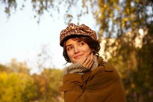 Portrait of positive beautiful brown-eyed young female with casual hairstyle leaning her chin on raised palm and smiling gently, wearing stylish clothes while posing over city garden photo