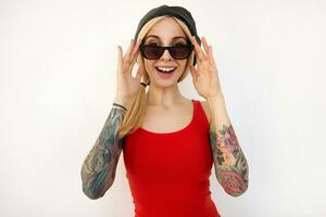 Cheerful young pretty tattooed blonde lady in elegant clothes keeping raised hand on her eyewear while looking gladly at camera with wide smile, isolated over white background photo