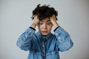 Discouraged young curly brunette female with natural makeup rumpling her hair with hands and looking distractedly in front of herself, wearing blue jeans coat over white background photo