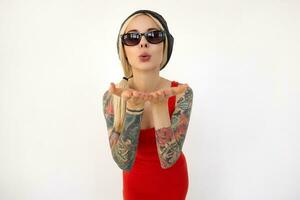 Indoor shot of young elegant tattooed blonde woman keeping her palms raised and pursing lips while sending air kiss to camera, posing over white background photo