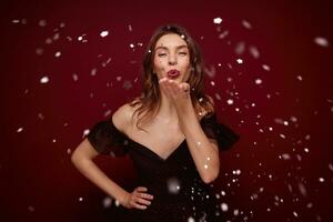 Studio photo of young brunette female dressed in elegant dress enjoying new year theme party while posing over claret background, raising palm and blowing silver confetti to camera