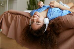 Young happy curly african american woman lying on the bed with her head down, broadly smiling and looks enjoying, listening favorite music in headphones. photo