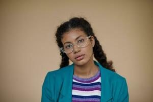 Stylish young curly dark skinned female with braids wearing eyewear while looking at camera with calm face, dressed in turquoise blazer and striped sweater while posing over beige background photo