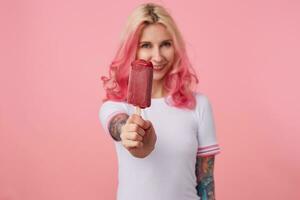 Portrait of happy cute girl with pink hair and tattooed hands, looking at the camera and offers to try delicious ice cream, smiling, standing over pink background, wearing a white t-shirt. photo