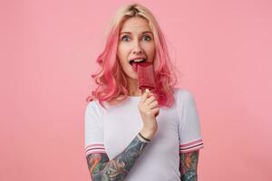 Photo of beautiful girl with pink hair and tattooed hands, wearing a white t-shirt, looking at the camera and trying to bite off the ice cream, standing over pink background.