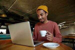 Shot of young dark skinned male with beard sitting at table in city cafe and drinking cup of tea, having lunch break and checking mails while waiting for order, wearing pink sweater and mustard cap photo