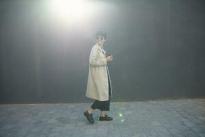 Outdoor photo of young curly stylish hipster female walking in city against black urban wall on warm sunny day, keeping cup of coffee in raised hand, dressed in black culottes and beige trench coat