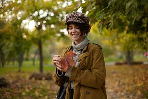 Good looking young cheerful brown haired woman with casual hairstyle holding yellowed leaf in her hands and smiling widely while waiting for her friends in city garden photo