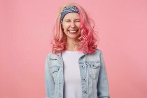 Happy cute smiling lady with pink hair and tattooed hands, standing over pink background with closed eyes, broadly smiling, wearing a white t-shirt and denim jacket. photo