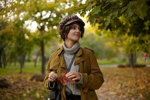 Fashionable young brown haired lady with bob hairstyle wearing trendy warm clothes while walking through yellow trees on warm autumn day, keeping leaf in raised hands and smiling positively photo