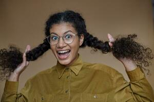 Overjoyed young curly brunette female with dark skin in glasses looking to camera with wide eyes and mouth opened, holding braids in raised hands while posing over beige background photo