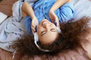 joven positivo Rizado mulato niña acostado en el cama con su cabeza abajo y cerrado ojos, escuchando favorito música en auriculares, en general sonriente y mira contento. foto