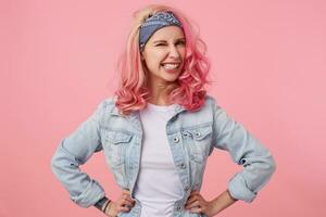 Portrait of happy cute girl with pink hair and tattooed hands, looking at the caera and winking, smiling and standing over pink background, wearing a white t-shirt and denim jacket. photo