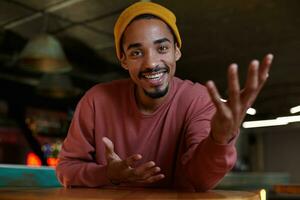 Portrait of charming positive brown eyed young bearded male with dark skin sitting over bar interior in pink sweater and mustard cap headdress, looking at camera with joyful smile and raising palm up photo