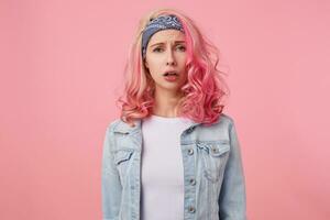 Close up of tired cute lady with pink hair, sad looking at the camera, can not find the right dress for the upcoming party. Standing over pink background, wearing a white t-shirt and denim jacket. photo