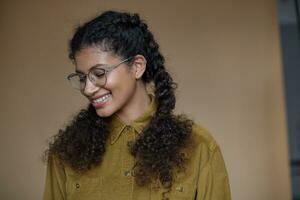 Indoor shot of good looking positive young dark skinned woman in glasses wearing her brown curly hair braided, dressed in mustard shirt while posing over beige background photo