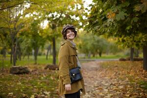 Stylish young happy lovely short haired brunette lady with bob hairstyle looking back at camera and smiling cheerfully while walking through park on warm autumn day photo