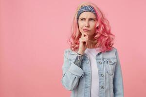 joven pensando bonito mujer con rosado cabello, soportes terminado rosado antecedentes Copiar espacio, mira arriba pensativamente y toques el mejilla con su dedo, usa un blanco camiseta y mezclilla chaqueta. foto