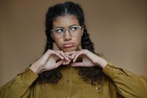 Sad dark skinned curly woman with braided hairstyle wearing glasses and holding chin on raised hands, looking aside with upset face and folding lips, isolated over beige background photo