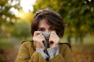 Outdoor photo of attractive brown-eyed young brunette female with natural makeup wearing stylish clothes while posing over park background and hiding her face