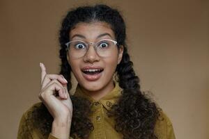 Portrait of lovely open-eyed young dark skinned lady with curly brown braided hair wearing glasses while posing over beige background, rounding her eyes and raising forefinger in having idea sign photo