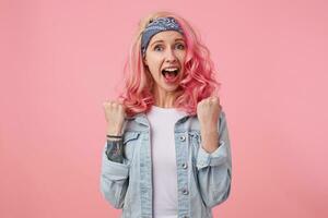 Happy lady with pink hair and tattooed hand, standing over pink background, wearing a white t-shirt and denim jacket. Screaming and celebrates victory of favorite football team with raising fists. photo