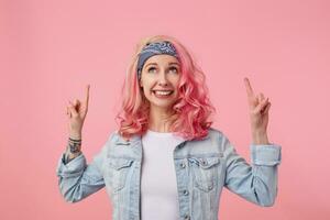 Happy cute smiling lady with pink hair and tattooed hands, standing over pink background, wearing a white t-shirt and denim jacket. looks up and points fingers at copy space above her head. photo