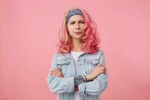 Frowning lady with pink hair and tattooed hand, looking at the camera with disapproval and discontent, standing over pink background with crossed arms, wearing a white t-shirt and denim jacket. photo