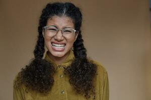 Portrait of unhappy young dark skinned lady wearing her curly brown hair braided, showing her perfect white teeth while crying sadly, dressed in casual wear while posing over beige background photo