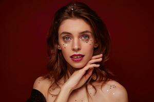 Studio photo of charming young brunette lady with burgundy lips and little silver stars on her face touching chin gently while looking at camera, isolated over claret background