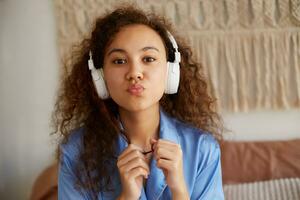 Close up of cute curly african american young woman listening favorite romantic music in headphones, sends kiss at the camera, looks happy. photo