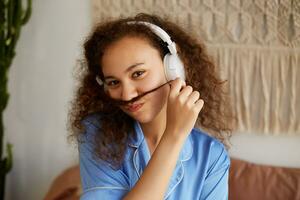 Portrait of young funny dark skinned woman with curly hair, makes a mustache from strands of hair, listening favorite song on headphones and feels good. photo