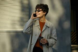 Pleasant looking cheerful young curly woman with casual hairstyle walking outside on sunny warm day, having pleasant talk on phone, wearing trendy clothes and stylish sunglasses photo