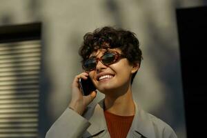 Close-up of beautiful young brunette curly female with charming smile holding mobile phone in raised hand, posing over urban environment in stylish clothes and vintage eyewear photo