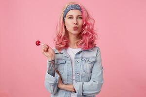 Young happy beautiful pink haired lady in denim shir, holding a lollipop, looks at the camera, sedns kiss, stands over pink background. photo